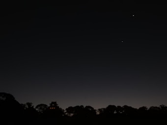 A dark night sky with subtle gradients, featuring a silhouette of trees and a structure resembling a traditional building in the foreground. Two bright celestial objects, possibly stars or planets, are visible in the sky.