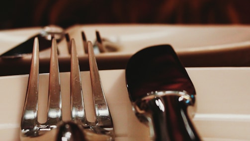 A close-up view of a dining table setting, featuring shiny metal cutlery including a fork and a knife placed on a white ceramic dish. The focus is on the reflective surface of the cutlery, with a soft depth of field blurring the background.