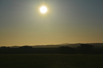 The image depicts a serene landscape with the sun setting or rising above a distant horizon, casting a gentle golden glow across the scene. Silhouettes of trees and hills create a peaceful and calm atmosphere, with a clear sky dominating the upper portion of the image.