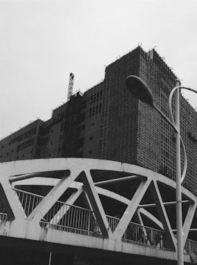 A black and white image of a construction site featuring a partially built multi-story building covered in scaffolding. In the foreground, a modern bridge with geometric designs and a street lamp are visible against a cloudy sky.