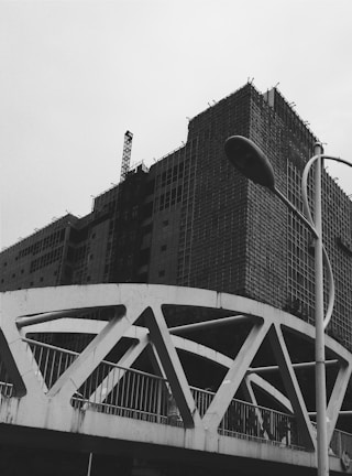 A black and white image of a construction site featuring a partially built multi-story building covered in scaffolding. In the foreground, a modern bridge with geometric designs and a street lamp are visible against a cloudy sky.
