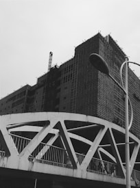 A black and white image of a construction site featuring a partially built multi-story building covered in scaffolding. In the foreground, a modern bridge with geometric designs and a street lamp are visible against a cloudy sky.
