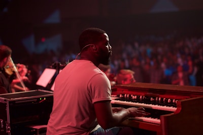 A musician plays a keyboard in a dimly lit concert setting. The background is filled with a blurred audience, creating a sense of a live performance. The musician is focused on playing, with sheet music in the background and another musician visible playing a string instrument.