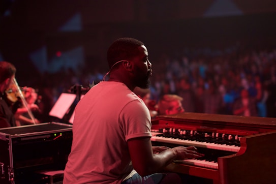 A musician plays a keyboard in a dimly lit concert setting. The background is filled with a blurred audience, creating a sense of a live performance. The musician is focused on playing, with sheet music in the background and another musician visible playing a string instrument.