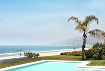 A picturesque coastal scene featuring a pristine swimming pool in the foreground, bordered by well-maintained grass and a lone palm tree. Beyond the pool, a sandy beach stretches alongside the shimmering blue ocean, with gentle waves and distant hills in the background under a clear sky.