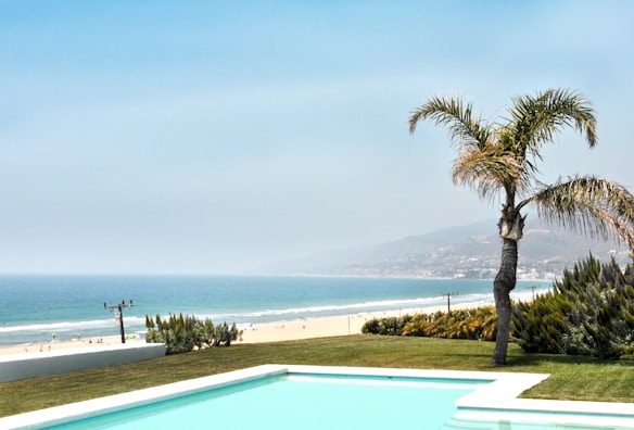 A picturesque coastal scene featuring a pristine swimming pool in the foreground, bordered by well-maintained grass and a lone palm tree. Beyond the pool, a sandy beach stretches alongside the shimmering blue ocean, with gentle waves and distant hills in the background under a clear sky.