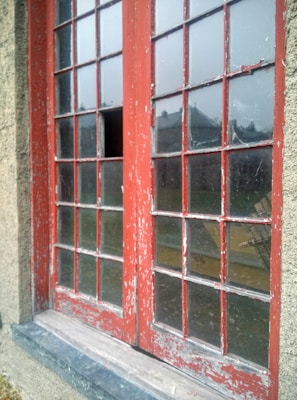 A weathered, red-painted wooden window set in a stone or concrete wall. The window features a grid of small glass panes, some of which are broken or missing, revealing the outdoors. The paint is peeling, giving a sense of age and neglect. Through the glass, a blurry reflection of buildings and a dreary sky is visible.
