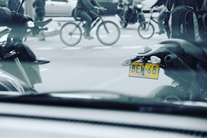 Multiple motorcycles and bicycles are seen in an urban setting. A visible license plate on a motorcycle reads 'BEN 68' from Colombia. The image displays movement and activity as people ride through the street.