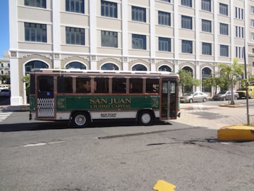 A small green and white city bus labeled 'San Juan Ciudad Capital' is parked on a street. The background features a large, white building with multiple windows and palm trees along the sidewalk. The street is mostly empty, with a few cars parked nearby.