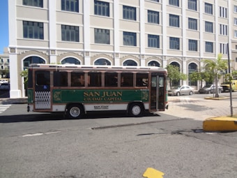 A small green and white city bus labeled 'San Juan Ciudad Capital' is parked on a street. The background features a large, white building with multiple windows and palm trees along the sidewalk. The street is mostly empty, with a few cars parked nearby.