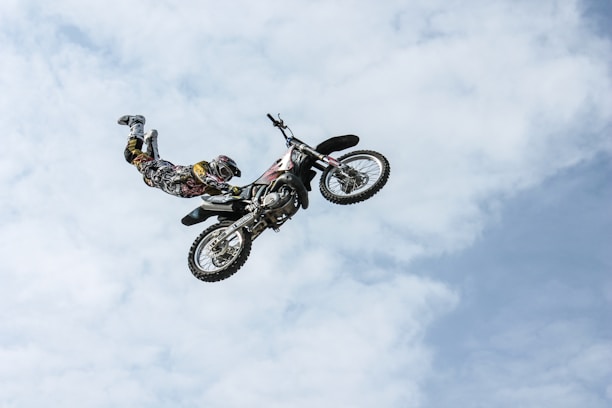 A motorcyclist performs an aerial stunt on a motocross bike against a backdrop of a partly cloudy sky. The rider is mid-air, extended away from the bike in a maneuver that showcases skill and balance.