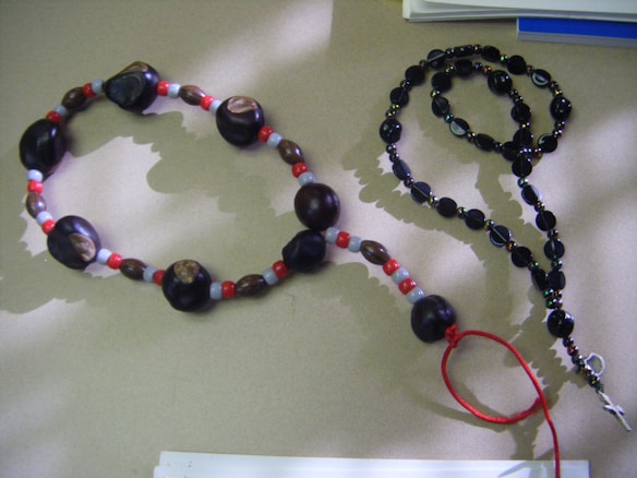 Two sets of beads are arranged on a light brown surface. One set consists of large, dark wooden beads interspersed with smaller red and light blue beads, forming a loop with a red string at the end. The other set is a black beaded rosary with a small metal cross attached.