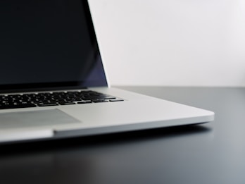 A sleek, silver laptop with a black keyboard sits on a dark gray surface against a white wall. The laptop's screen is turned off, and the angle is captured from the bottom corner, showcasing its minimalist design.