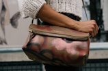 A person is holding a stylish handbag with a floral pattern in shades of brown, pink, and black. The person is wearing a white knitted sweater. The background shows a partially visible building and mural featuring a large butterfly.