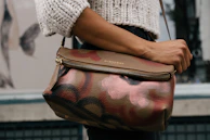 A person is holding a stylish handbag with a floral pattern in shades of brown, pink, and black. The person is wearing a white knitted sweater. The background shows a partially visible building and mural featuring a large butterfly.