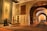 An elegantly decorated hall with arched brick ceilings and walls showcases various intricately patterned rugs and carpets. A decorative bench with ornate metalwork sits on top of a geometric-patterned carpet. The lighting highlights the textures and designs of the textiles, creating a sense of luxury and sophistication.