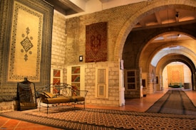 An elegantly decorated hall with arched brick ceilings and walls showcases various intricately patterned rugs and carpets. A decorative bench with ornate metalwork sits on top of a geometric-patterned carpet. The lighting highlights the textures and designs of the textiles, creating a sense of luxury and sophistication.
