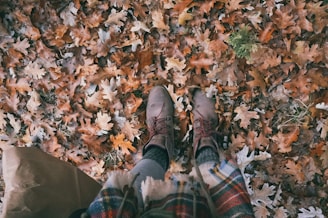 A pair of brown boots standing on a bed of fallen autumn leaves, displaying various shades of orange and brown. The person is wearing cozy, patterned clothing that suggests a cool weather setting. The leaves are scattered densely, with some green foliage peeking through.