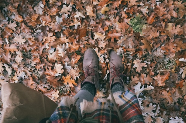A pair of brown boots standing on a bed of fallen autumn leaves, displaying various shades of orange and brown. The person is wearing cozy, patterned clothing that suggests a cool weather setting. The leaves are scattered densely, with some green foliage peeking through.