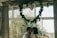 A couple is standing close together, sharing an intimate moment in a well-lit room with large windows. Above them, a heart-shaped arrangement of greenery and a metallic heart decoration hang from the ceiling. The room is decorated modestly, with curtains flanking the windows, and a lush garden is visible outside.