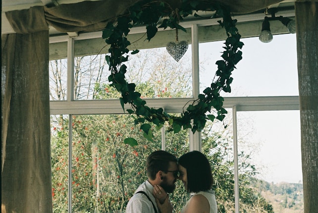A couple is standing close together, sharing an intimate moment in a well-lit room with large windows. Above them, a heart-shaped arrangement of greenery and a metallic heart decoration hang from the ceiling. The room is decorated modestly, with curtains flanking the windows, and a lush garden is visible outside.