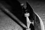 A close-up black and white photograph of a skateboard lying upside down on a textured surface, with the focus on the wheels and trucks. Intricate patterns are visible on the side of the skateboard deck.