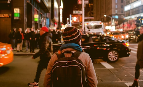 back view of woman wearing a backpack and beanie waiting to cross the road on a busy street at night in New York City, USA
