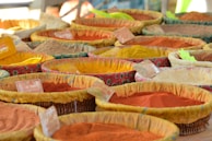 A variety of colorful spices displayed in woven baskets at a market. Each basket contains a different spice, with hues ranging from vibrant red and orange to dull brown and yellow. Small labels are attached to the baskets, presumably indicating the names or prices of the spices. The fabric lining the baskets is patterned and colorful, complementing the spices.