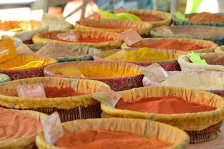 A variety of colorful spices displayed in woven baskets at a market. Each basket contains a different spice, with hues ranging from vibrant red and orange to dull brown and yellow. Small labels are attached to the baskets, presumably indicating the names or prices of the spices. The fabric lining the baskets is patterned and colorful, complementing the spices.