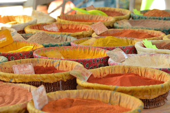 A variety of colorful spices displayed in woven baskets at a market. Each basket contains a different spice, with hues ranging from vibrant red and orange to dull brown and yellow. Small labels are attached to the baskets, presumably indicating the names or prices of the spices. The fabric lining the baskets is patterned and colorful, complementing the spices.