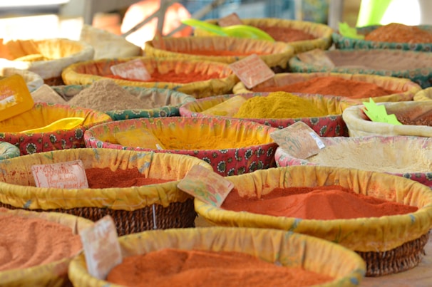 A variety of colorful spices displayed in woven baskets at a market. Each basket contains a different spice, with hues ranging from vibrant red and orange to dull brown and yellow. Small labels are attached to the baskets, presumably indicating the names or prices of the spices. The fabric lining the baskets is patterned and colorful, complementing the spices.