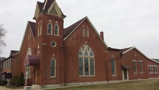 A large brick building with Gothic architectural elements, featuring arched windows and a prominent tower with a spire. The windows contain stained glass, and there is a maroon awning above the main entrance. The building is surrounded by a grass lawn and a sidewalk curves around the front.