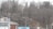 A landscape featuring a winding road flanked by leafless trees. In the foreground, there's a small building with a sign reading 'U-Go Auto Sales CARS.' The background consists of densely packed trees on a hillside with a few houses visible.