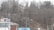 A landscape featuring a winding road flanked by leafless trees. In the foreground, there's a small building with a sign reading 'U-Go Auto Sales CARS.' The background consists of densely packed trees on a hillside with a few houses visible.