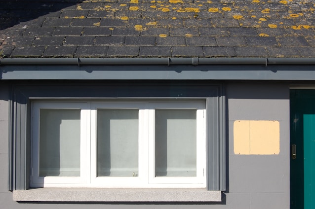 A small section of a building facade featuring a rectangular white-framed window with three panes. Above, grey slate roof tiles with patches of yellow lichen can be seen. The building wall is painted gray, and there is a small beige panel next to the window.