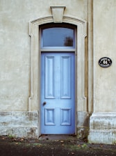A wooden door painted in light blue is set within an ornate stone archway. The surrounding wall is made of a textured beige material. The door features decorative paneling and is topped with a semi-circular window. To the right, a round black sign displays the address 'SUSSEX ST. 64 LINTON'. Fallen leaves are scattered on the ground near the base of the door.