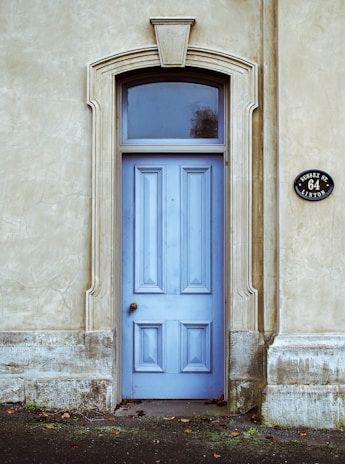 A wooden door painted in light blue is set within an ornate stone archway. The surrounding wall is made of a textured beige material. The door features decorative paneling and is topped with a semi-circular window. To the right, a round black sign displays the address 'SUSSEX ST. 64 LINTON'. Fallen leaves are scattered on the ground near the base of the door.