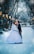A couple dressed in formal wedding attire shares a kiss in a snow-covered park. The scene is framed by tall trees with bare branches, and a row of glowing streetlamps lines the path. Snow gently falls around them, adding a magical touch to the scene.