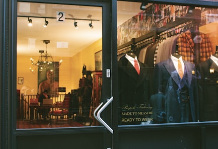 A storefront window of a bespoke tailoring shop displays mannequins dressed in formal suits. Inside, a cozy interior room is visible with a warm yellow light, featuring a person at a desk with a laptop, decorative furniture, and artwork on the walls.