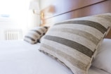 A neatly made bed with a wooden headboard is partially visible. Two large pillows with striped patterns in shades of beige and gray are prominently placed against the headboard. A small wall-mounted lamp is visible in the background, along with a bright window allowing natural light to illuminate the room. The overall setting appears clean, modern, and minimalistic.