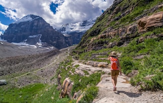 A person wearing a hat and carrying a red backpack is hiking along a rocky mountain trail with patches of greenery. Majestic snow-capped mountains rise in the background under a partly cloudy sky.