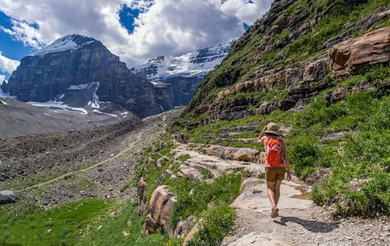 A person wearing a hat and carrying a red backpack is hiking along a rocky mountain trail with patches of greenery. Majestic snow-capped mountains rise in the background under a partly cloudy sky.