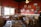 A cozy restaurant interior with wooden chairs and tables covered with checkered tablecloths. The red walls are adorned with various framed black-and-white photographs and pictures. A shelf above the window holds an array of blue kitchenware. Condiments are neatly placed on the tables, and sunlight filters through the large window.