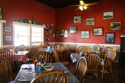 A cozy restaurant interior with wooden chairs and tables covered with checkered tablecloths. The red walls are adorned with various framed black-and-white photographs and pictures. A shelf above the window holds an array of blue kitchenware. Condiments are neatly placed on the tables, and sunlight filters through the large window.