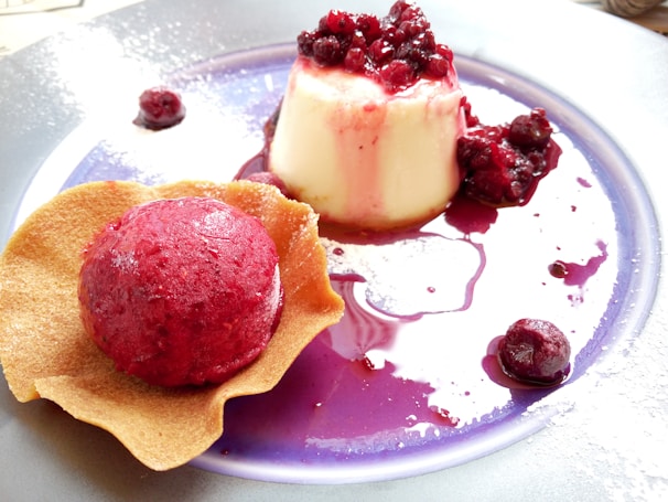 A dessert plate features a creamy panna cotta topped with red berries and a scoop of raspberry sorbet beside a crisp wafer. The sauce from the berries spreads artistically across the plate, creating an inviting presentation.
