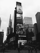A bustling urban scene featuring towering skyscrapers with numerous large, illuminated advertisements. Prominent brands such as Gree, Coca-Cola, and Hyundai are displayed on the billboards. The area is crowded with people walking and engaging in various activities. The photo is in black and white, giving it a classic, timeless feel.