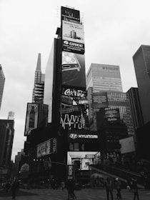 A bustling urban scene featuring towering skyscrapers with numerous large, illuminated advertisements. Prominent brands such as Gree, Coca-Cola, and Hyundai are displayed on the billboards. The area is crowded with people walking and engaging in various activities. The photo is in black and white, giving it a classic, timeless feel.