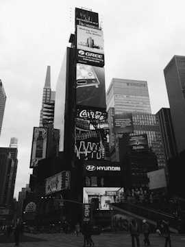 A bustling urban scene featuring towering skyscrapers with numerous large, illuminated advertisements. Prominent brands such as Gree, Coca-Cola, and Hyundai are displayed on the billboards. The area is crowded with people walking and engaging in various activities. The photo is in black and white, giving it a classic, timeless feel.