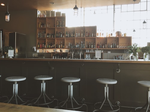 A modern bar interior with a row of metal stools lined up against a wooden bar counter. Shelves behind the bar are stocked with a variety of liquor bottles. Glasses and bar tools are neatly arranged. The space is well-lit by large windows and industrial-style pendant lights hang from the ceiling.