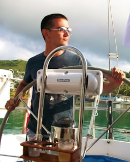A person wearing sunglasses is steering a boat, with lush green hills and cloudy skies in the background. The boat features a steering wheel and navigation equipment branded 'NavPod'. There are glasses with drinks on a wooden holder attached to the equipment.